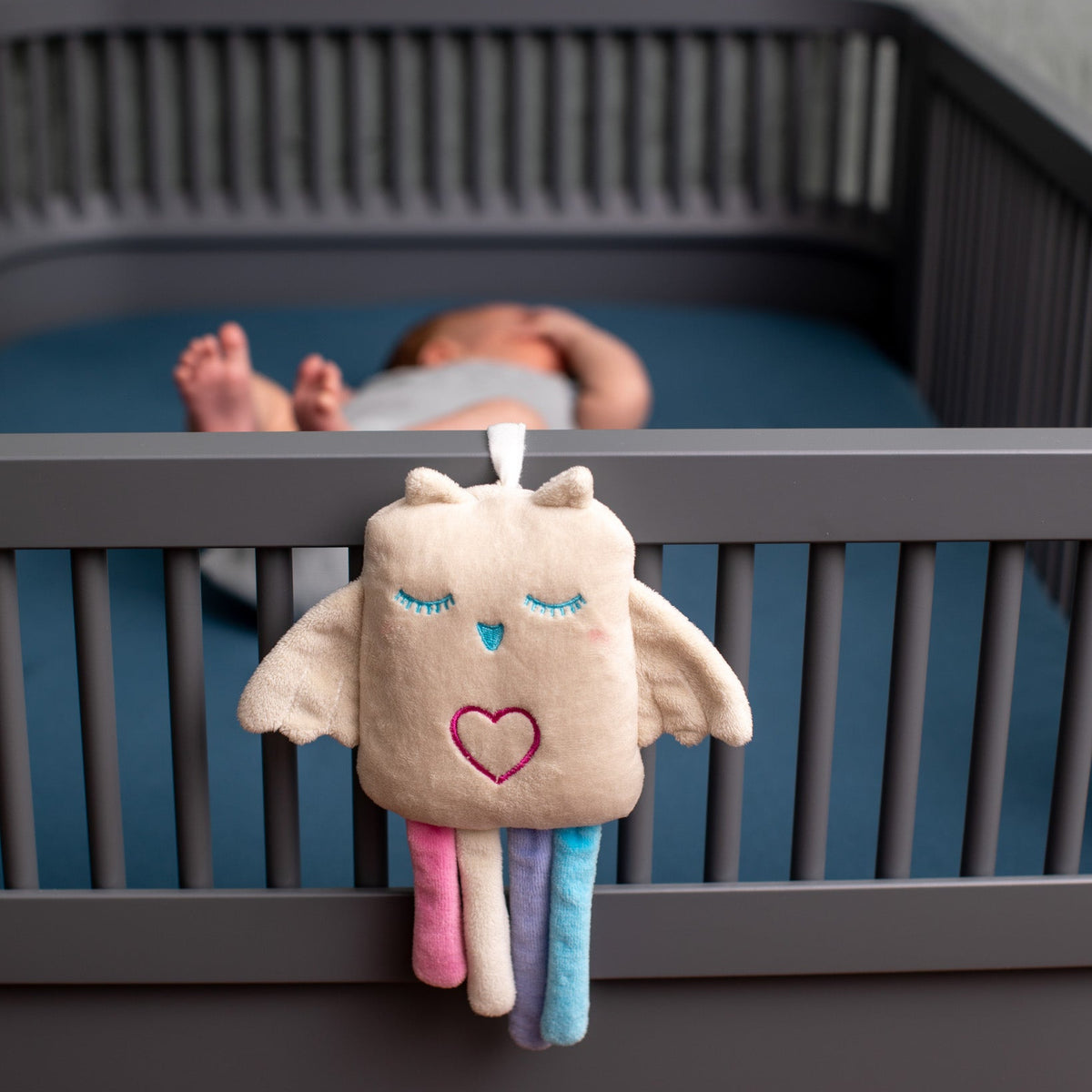 A plush baby toy shaped like a sleeping angel with pastel wings and legs hangs from the front rail of a dark gray crib. A baby is lying in the crib in the blurred background.